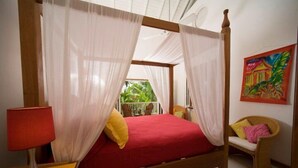A four-poster bed with sheer curtains, a red bedspread, and a red lamp on a wooden nightstand. - Tobago Hibiscus Villas and Apartments (Bethel)