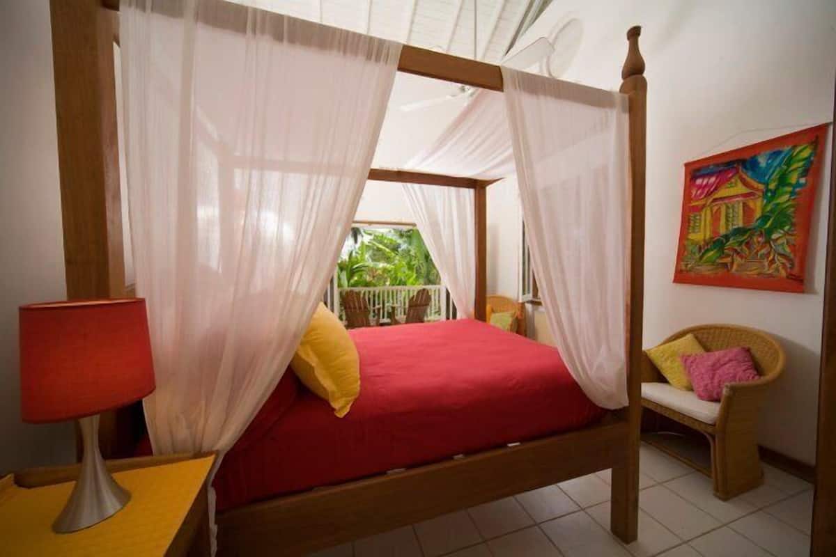 Un lit à baldaquin avec des rideaux légers, une couvre-lit rouge et une lampe rouge posée sur une table de chevet en bois.