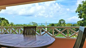 A round wooden table with chairs on a balcony with a white railing, overlooking a lush landscape and the ocean. - Tobago Hibiscus Villas and Apartments (Bethel)