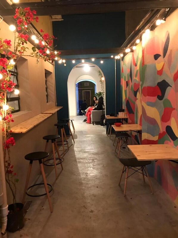 Un pasillo estrecho con mesas y sillas, un mural colorido en la pared y luces colgantes.