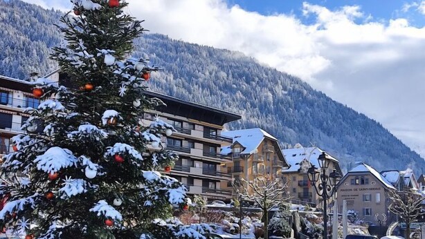 A snow-covered Christmas tree in a town square with buildings and mountains in the background.