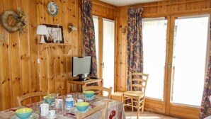 A wooden interior with a dining table set for four, a television on a stand, and a window with curtains.