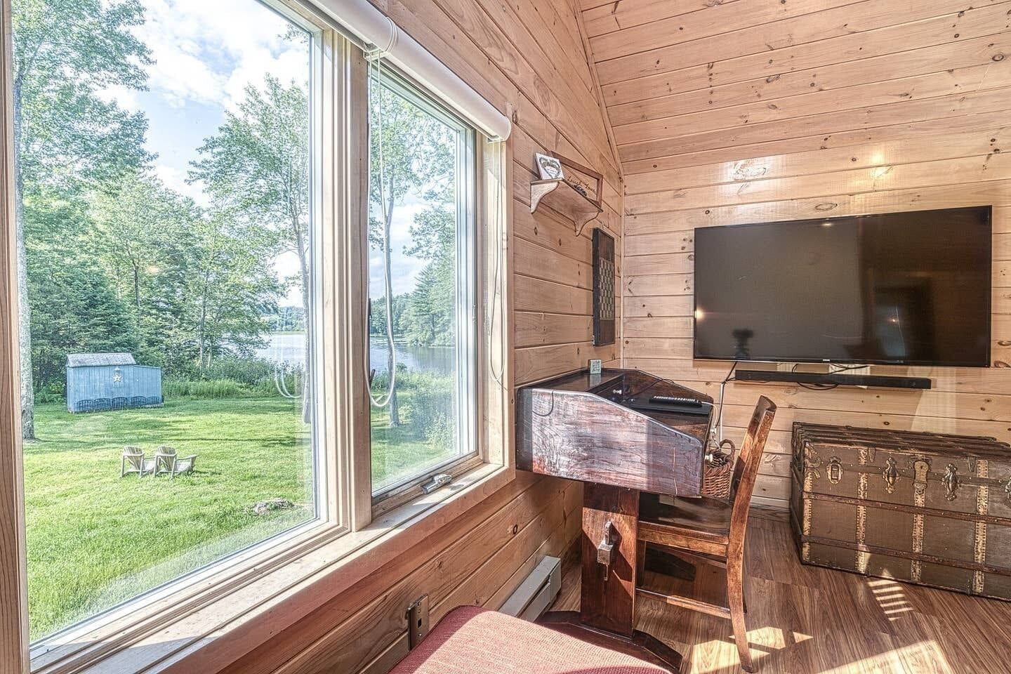 Una cabaña de madera con un ventanal grande, un televisor de pantalla plana, una mesa de madera y un cobertizo pequeño en el patio.