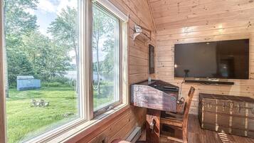 Una cabaña de madera con un ventanal grande, un televisor de pantalla plana, una mesa de madera y un cobertizo pequeño en el patio.
