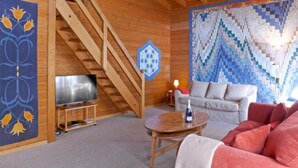 Un intérieur en bois avec un escalier, un canapé, un téléviseur et un mur orné d’un motif géométrique.