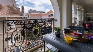 Un balcon orné d’une balustrade en fer forgé, une petite table avec un pot de fleurs, et une vue sur les bâtiments qui se dressent à l’arrière-plan.