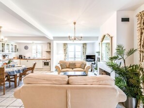 A spacious living area with a sofa, dining table, and chairs. There is a kitchen in the background with cabinets and a clock.