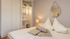 Ein ordentlich bezogenes Bett mit Kissen und Decke, eine Nachttischlampe, ein runder Spiegel an der Wand und ein Badezimmer mit Waschbecken und Handtuchhalter.