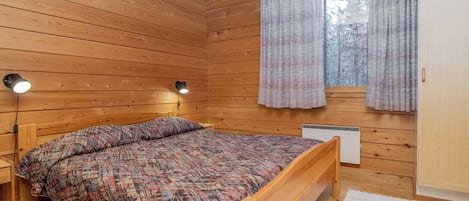 Dormitorio de cabaña de madera con cama, dos lámparas fijadas a la pared, una ventana con cortinas y un radiador.