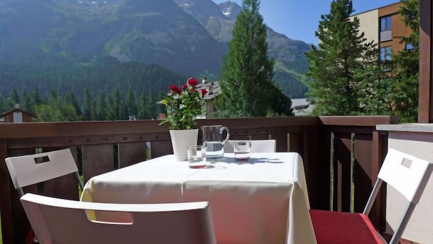 A table set outdoors with a mountain view, white chairs with red cushions, and a potted plant with red flowers.
