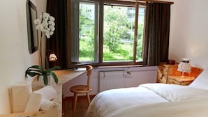 A bedroom with a bed, a desk, a chair, a radiator, a window with curtains, and a bookshelf.