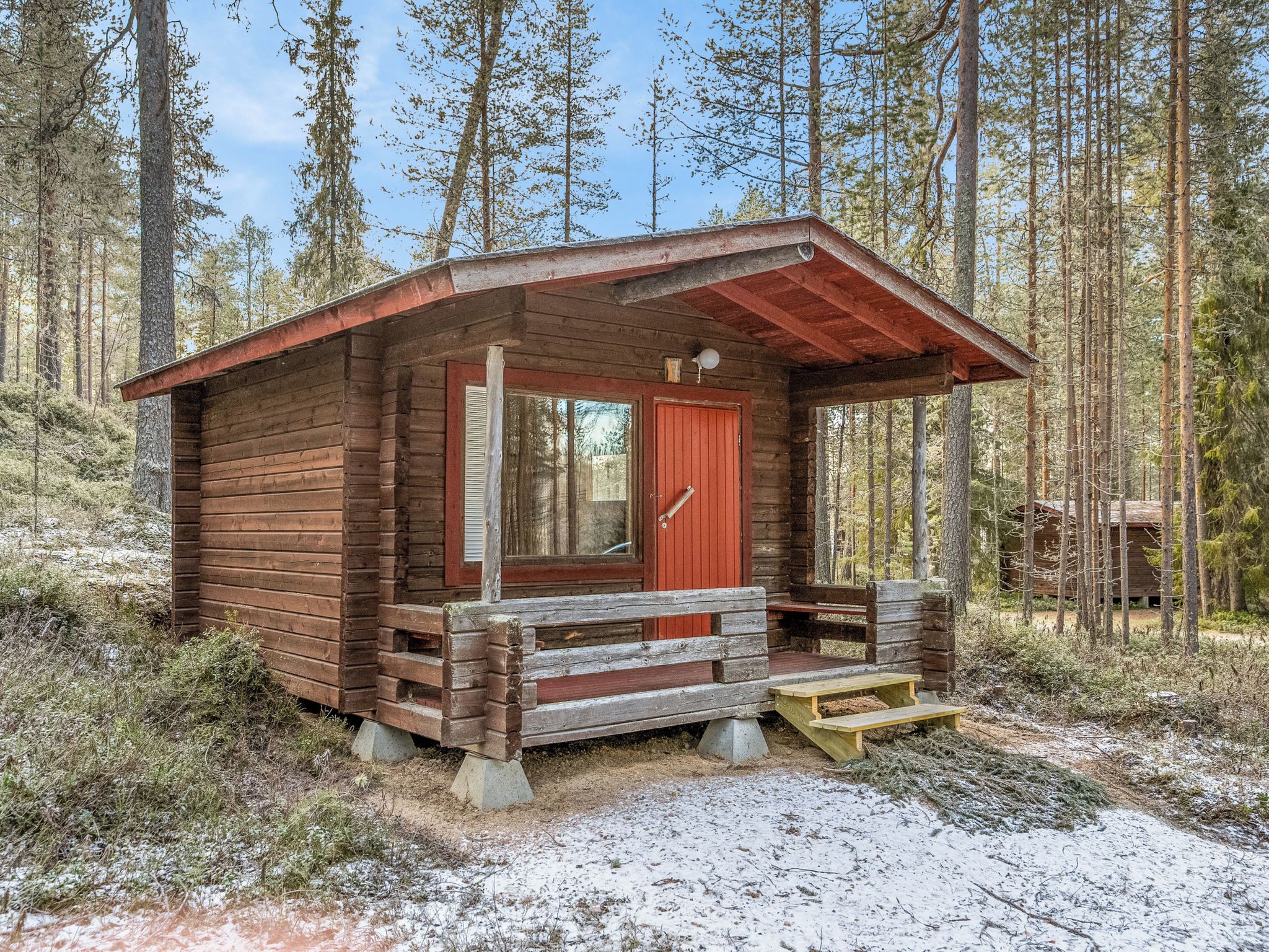 Una cabaña de madera pequeña con puerta roja y un porche, rodeada de un bosque.