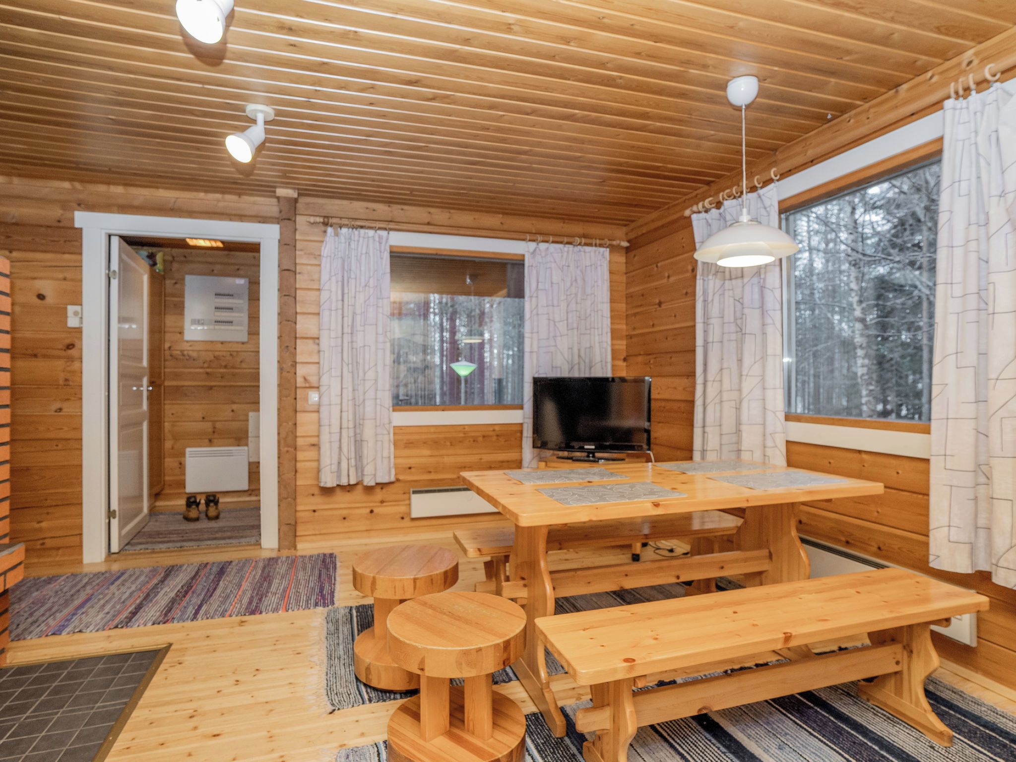 Interior de una cabaña de madera con una mesa de comedor, bancos, un televisor y ventanas grandes.