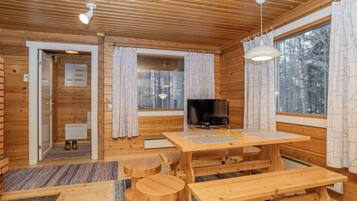 Interior de una cabaña de madera con una mesa de comedor, bancos, un televisor y ventanas grandes.