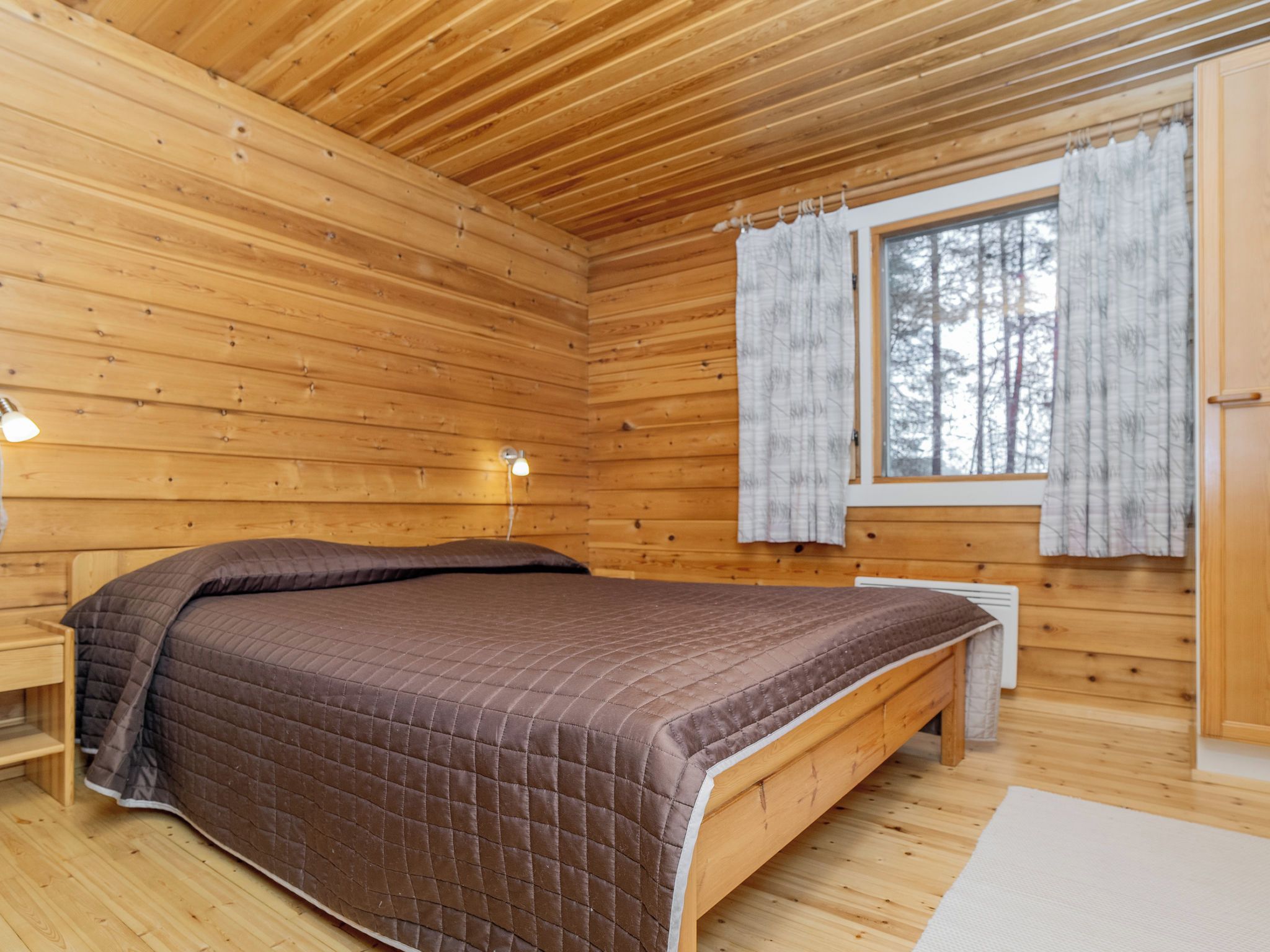 Un dormitorio de cabaña de madera con una cama, una mesita de noche, una ventana con cortinas y un calentador fijado a la pared.