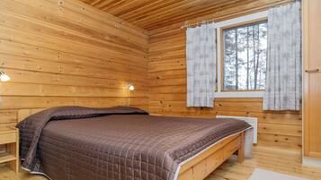Un dormitorio de cabaña de madera con una cama, una mesita de noche, una ventana con cortinas y un calentador fijado a la pared.