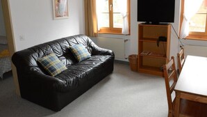 A black leather sofa with plaid cushions, a wooden TV stand, and a dining table in a room with white walls and a window with curtains.