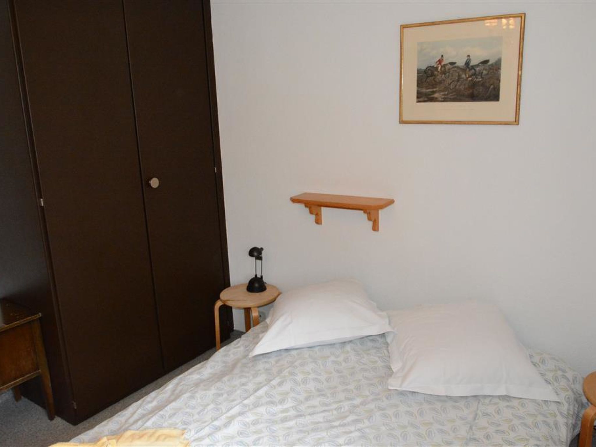 Un dormitori amb llit, taula de nit, làmpada, armari i un quadre amb marc a la paret.