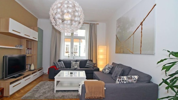 Ein modernes Wohnzimmer mit grauem Sofa, weißem Couchtisch und einer großen Chandelier. Ein Fernseher steht auf einem Holzgestell, und an der Wand hängt ein Gemälde.