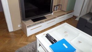 Ein Wohnzimmer mit Flachbildfernseher auf einer Holz-TV-Konsole, einem weißen Couchtisch mit Fernbedienung und einer blauen Ordner, sowie einem Bücherregal mit DVDs.