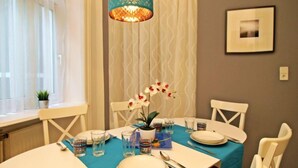 Ein Essbereich mit einem für vier Personen gedeckten Tisch, einer türkisfarbenen Tischdecke, weißen Stühlen, einer Vase mit weißen Blumen und einer dekorativen Leuchte.