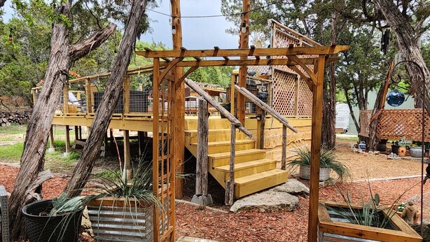 A wooden outdoor staircase leading up to a wooden deck, surrounded by trees and potted plants. - Home On The Range Charming Home - large private deck w/hot tub and Great Views (Wimberley)