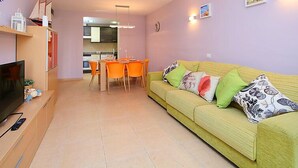 Un salon avec un canapé jaune, des fauteuils orange, un téléviseur et une cuisine en arrière-plan.