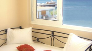 A bed with a metal frame, white bedding, and a red pillow. A window with a view of a building and water is visible. - Dorana Trekking Hotel (Karpathos)