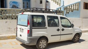 A white van parked on a street with a metal rack on the roof, a visible license plate, and a building with blue doors and white walls in the background. - Dorana Trekking Hotel (Karpathos)