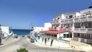 A coastal street with buildings, including a studio named "Studios Dorana," a red-roofed building, and a clear view of the sea. - Dorana Trekking Hotel (Karpathos)