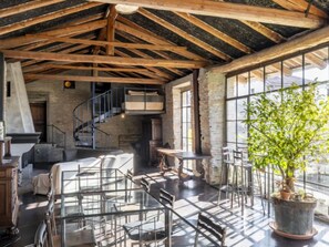 Une pièce spacieuse avec un plafond haut, des poutres apparentes, un escalier en colimaçon, une grande fenêtre et une plante en pot.