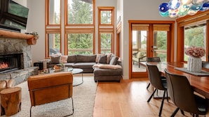 A spacious living room with a stone fireplace, large windows, a dining area, and a cozy seating arrangement. - 3BR | Taluswood | Ski-in/Ski-out | Private Hot Tub | Wood Burning Fireplace | Parking (Whistler)