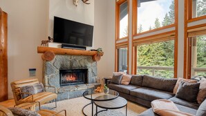 A cozy living room with a stone fireplace, a large TV mounted above it, and a comfortable seating area with a sofa and chairs. - 3BR | Taluswood | Ski-in/Ski-out | Private Hot Tub | Wood Burning Fireplace | Parking (Whistler)