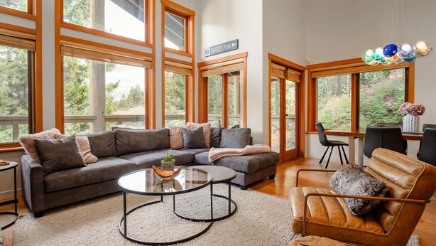 A spacious living room with a large gray sectional sofa, a round coffee table, and a leather armchair. Large windows provide a view of the surrounding forest. - 3BR | Taluswood | Ski-in/Ski-out | Private Hot Tub | Wood Burning Fireplace | Parking (Whistler)
