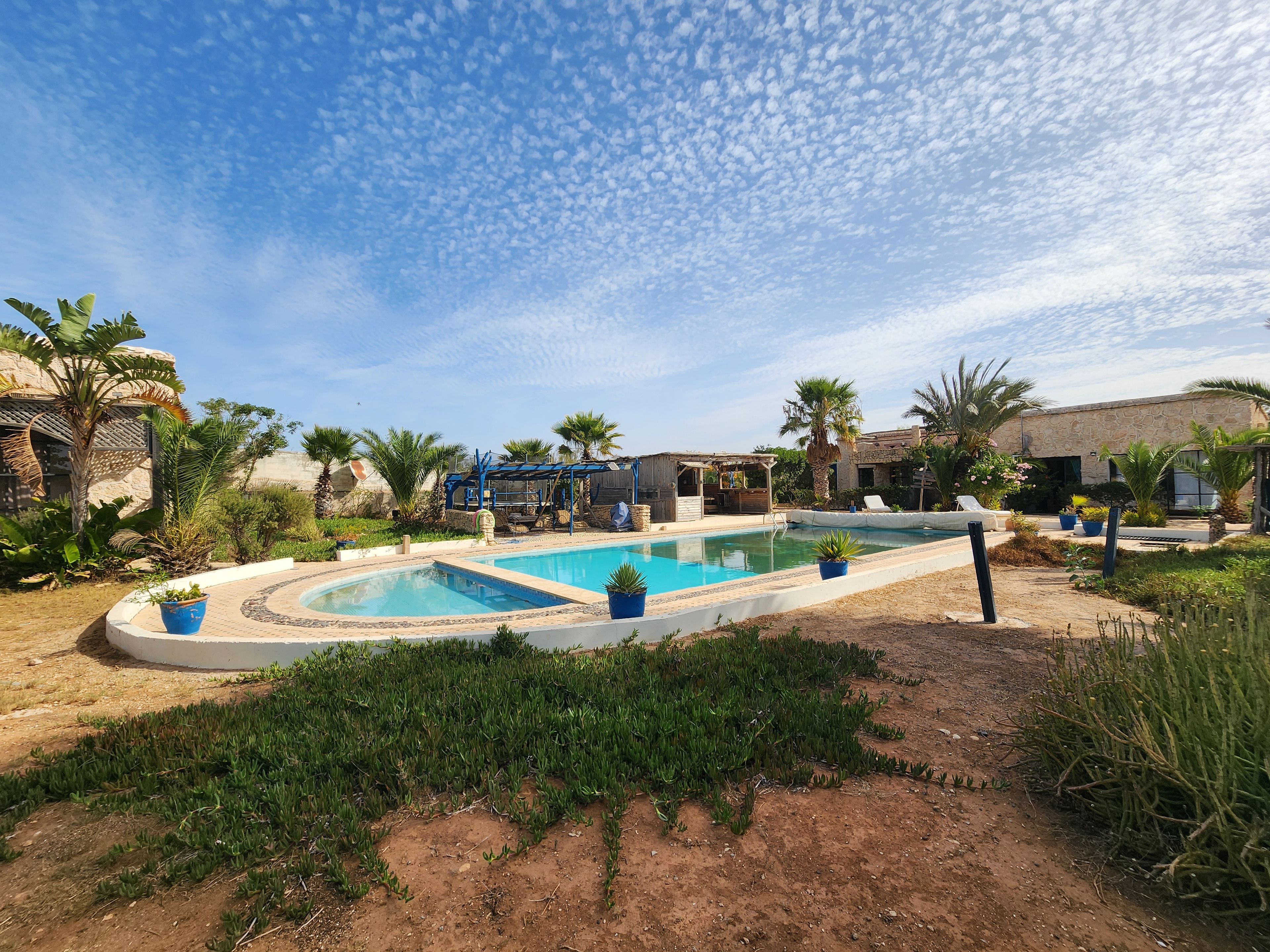 A swimming pool with a sunshade structure, surrounded by palm trees and a stone building.