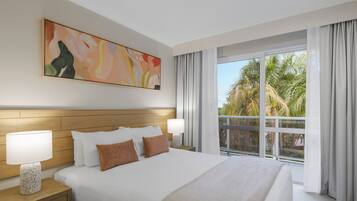A hotel room with a large bed, a nightstand with a lamp, a painting on the wall, and a view of palm trees through the window.