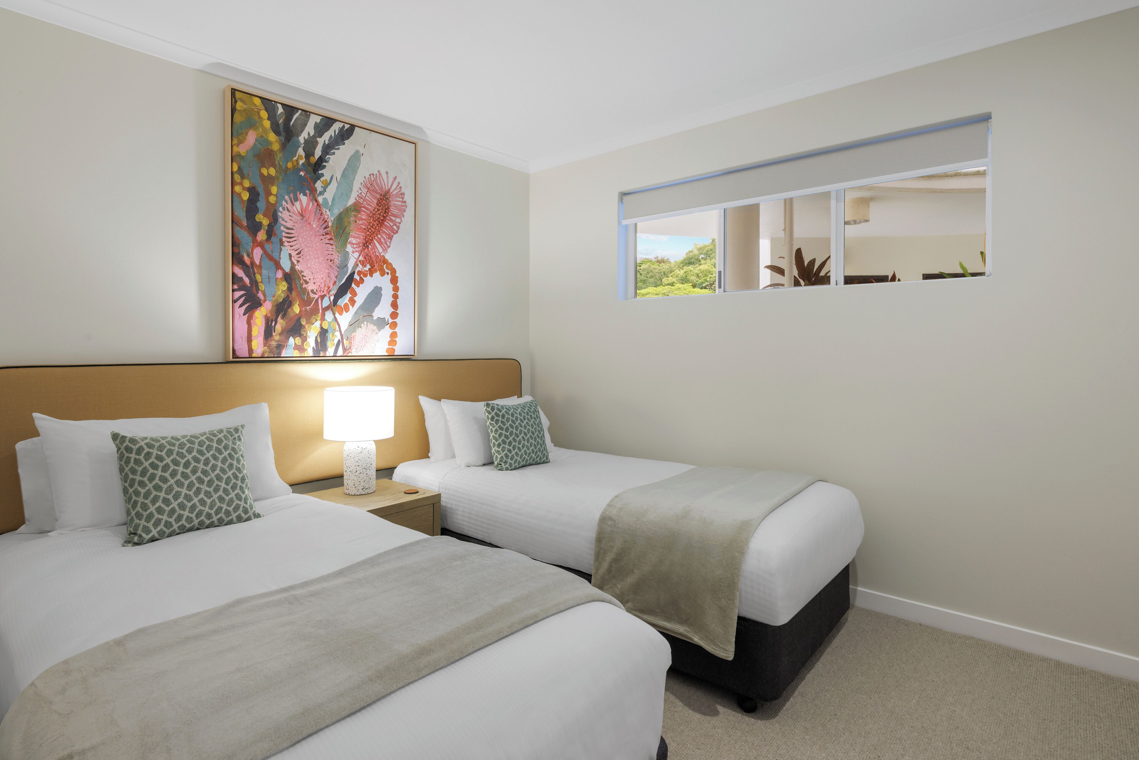 Hotelkamer met twee bedden, een nachtkastje met een lamp, een schilderij aan de muur en een raam met uitzicht op groen.