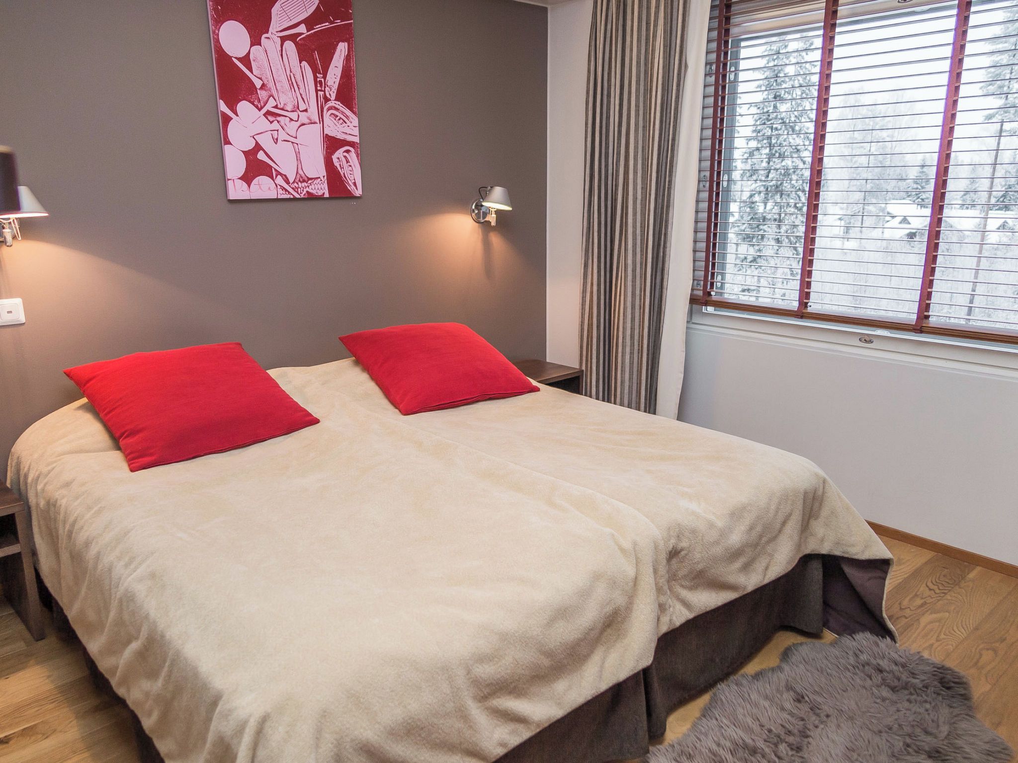 Una cama doble con almohadas rojas, una mesita de noche, una lámpara, una ventana con persianas y un cuadro en la pared.