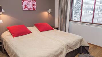 Un lit double avec des oreillers rouges, une table de chevet, une lampe, une fenêtre avec des stores et un tableau au mur.