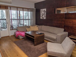 A modern living room with a sofa, a coffee table, and a chair. There is a large window with blinds, a fireplace, and a wall-mounted artwork. - Tahko spa suites orange a 9 by Interhome (Nilsiä)