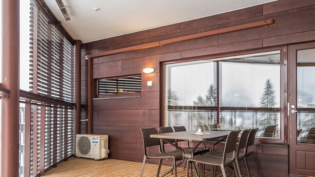 Een overdekt terras met een tafel en stoelen, een groot raam met uitzicht op bomen, en een airconditioning.