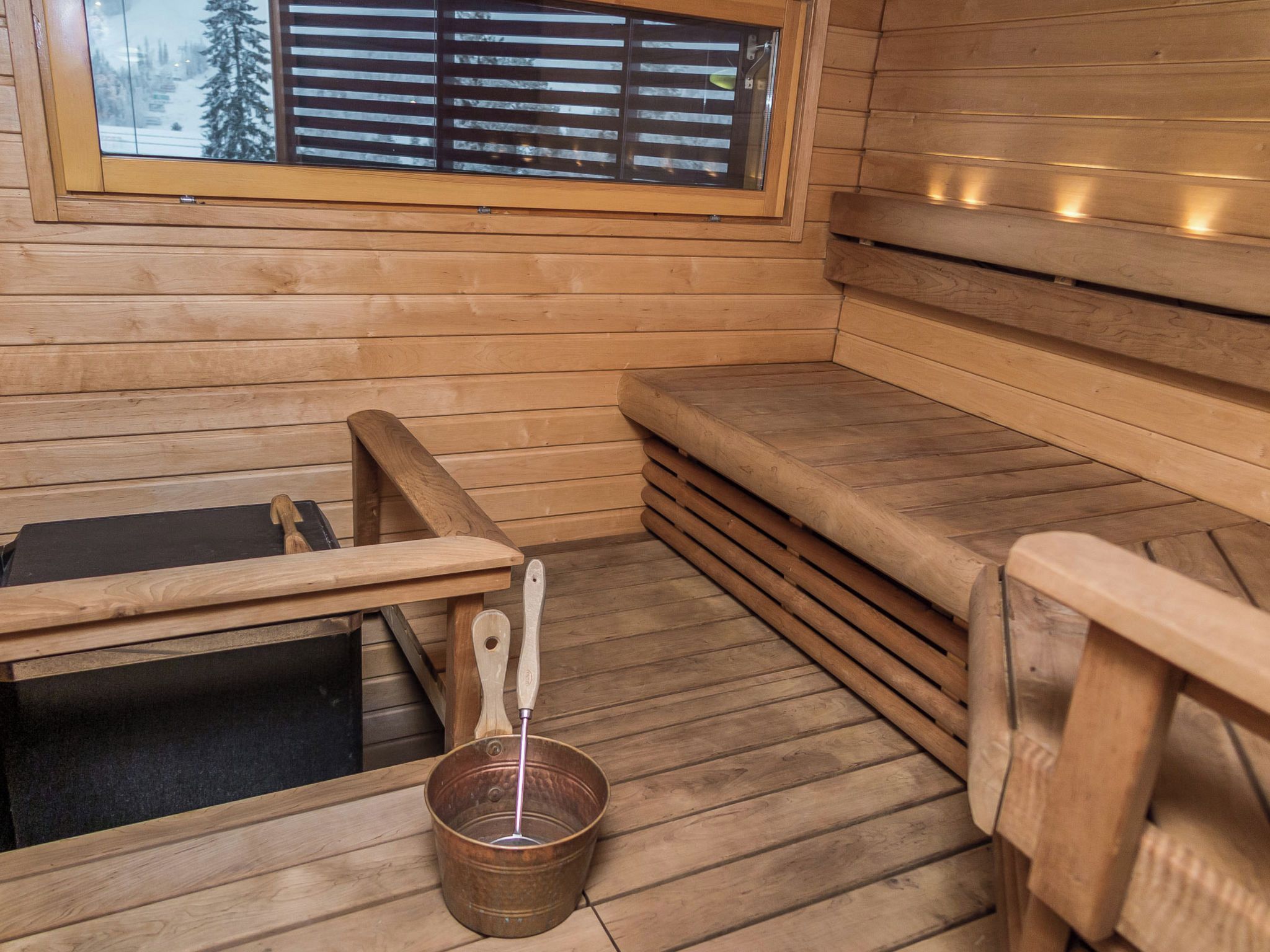 Un sauna en bois avec des bancs, un banc en bois, un seau en métal et une fenêtre avec des stores.