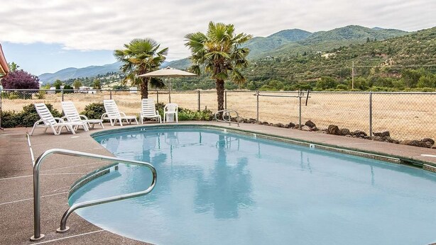 A swimming pool with lounge chairs and palm trees, surrounded by a fence and mountains in the background. - Ashland Valley Inn (Ashland)