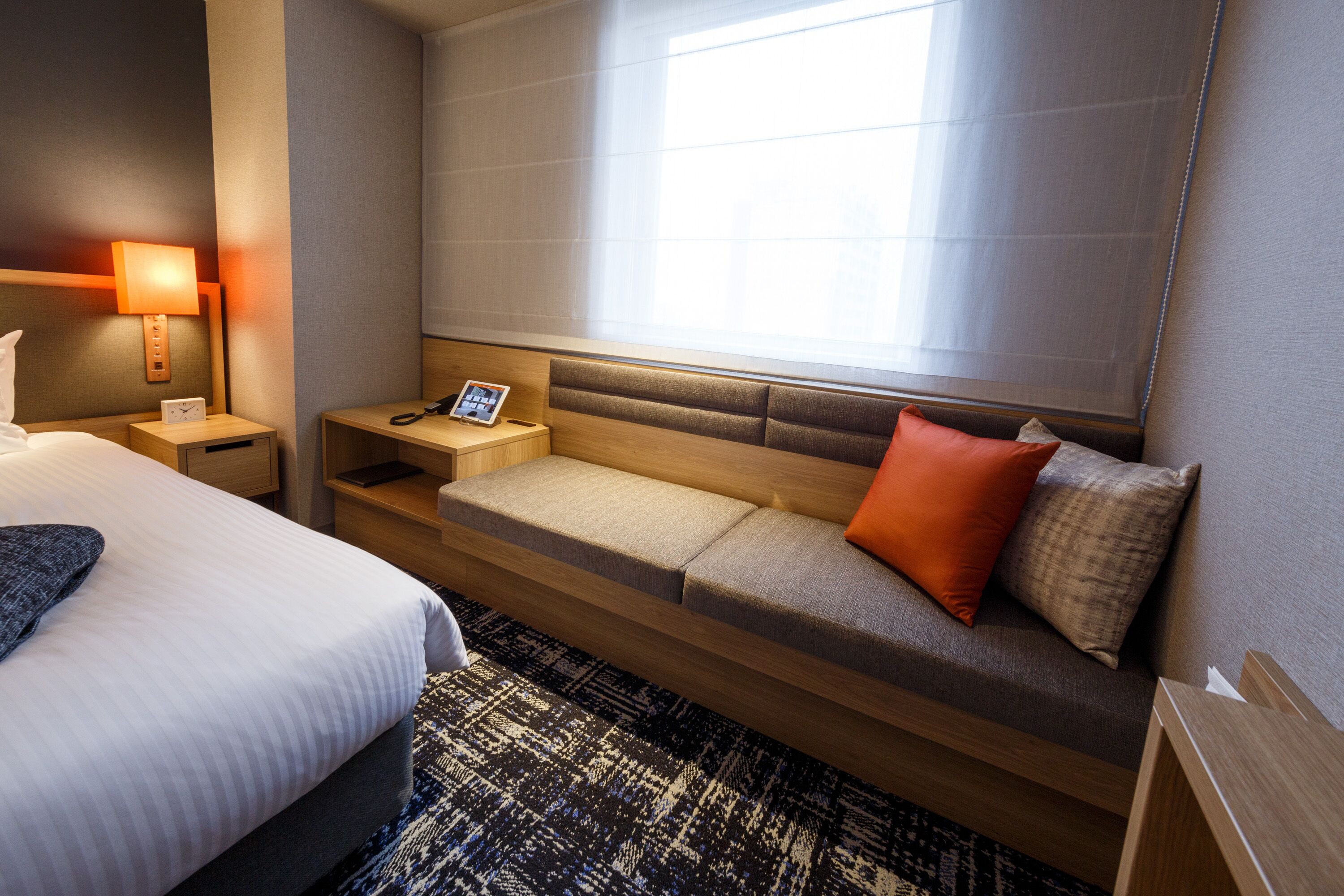 A hotel room with a large bed, a sofa with cushions, a bedside table with a lamp, and a window with blinds.