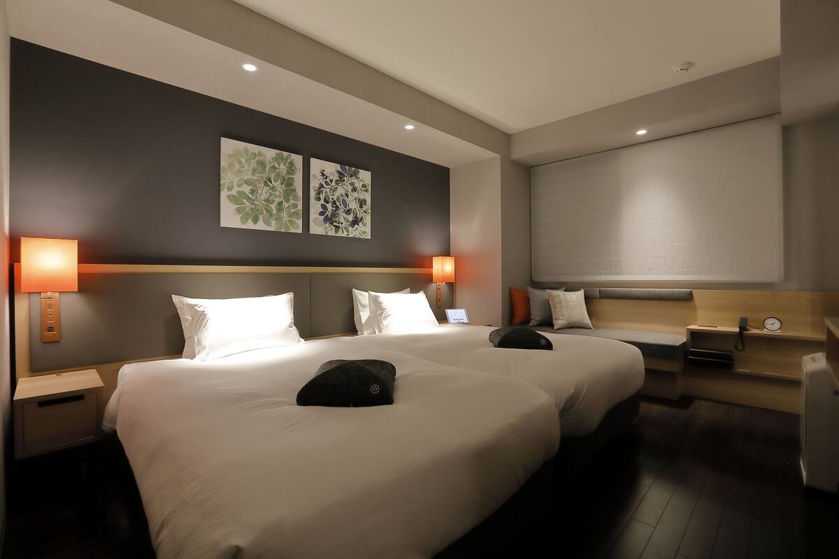 A hotel room with two beds, a bedside table with a lamp, a small desk with a chair, and a wall-mounted clock.