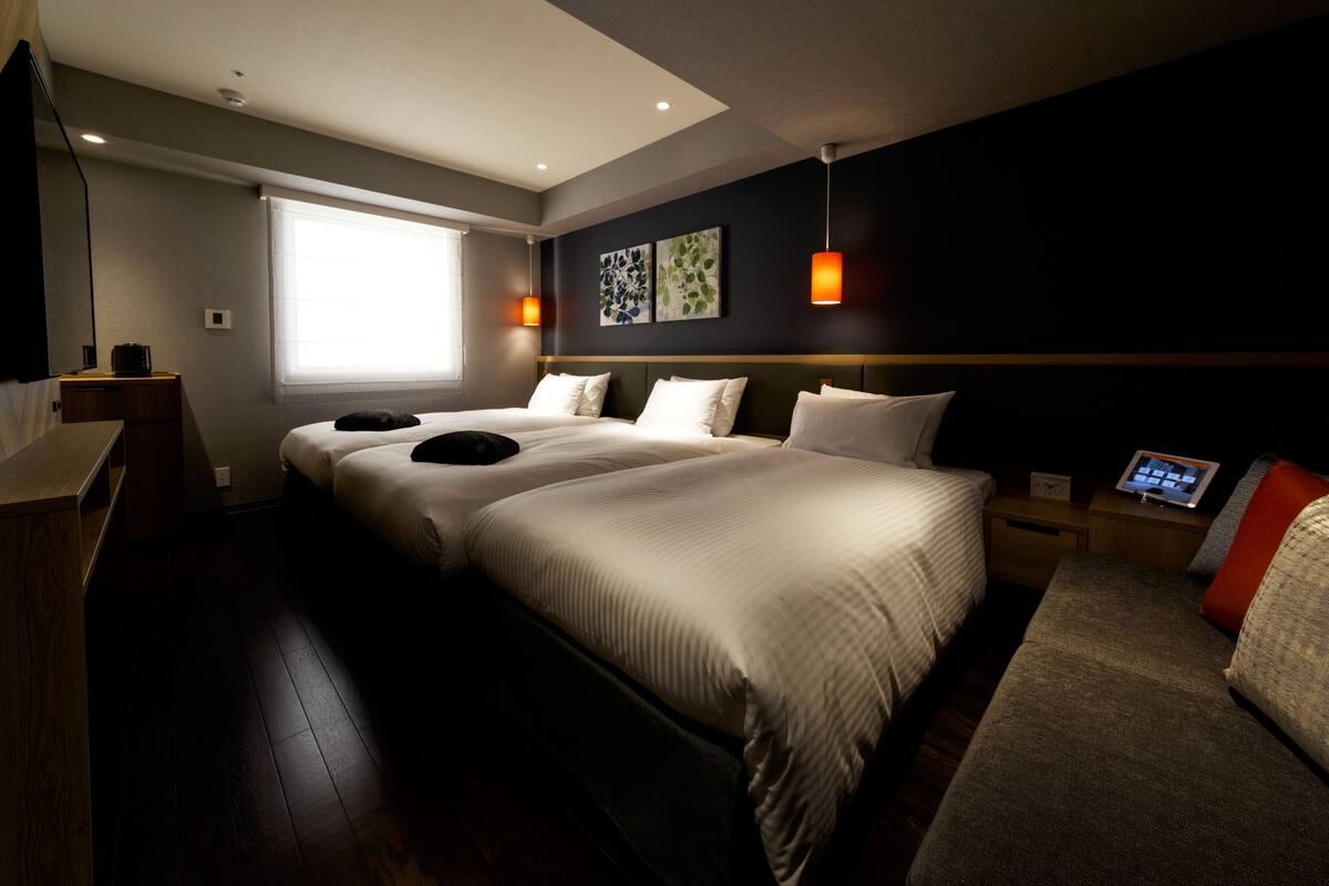 A hotel room with two beds, a bedside table with a lamp, a large window, and artwork on the wall.