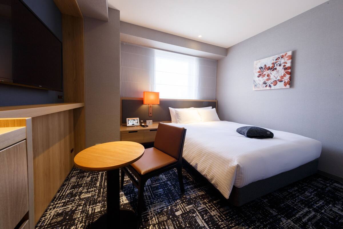 A hotel room with a bed, a desk, a chair, a lamp, and a painting on the wall.