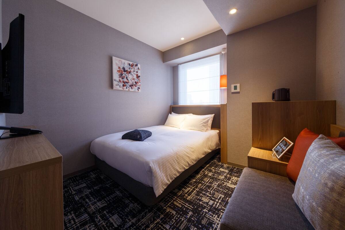 A hotel room with a bed, a desk, a television, a window, and a bedside table with a lamp.