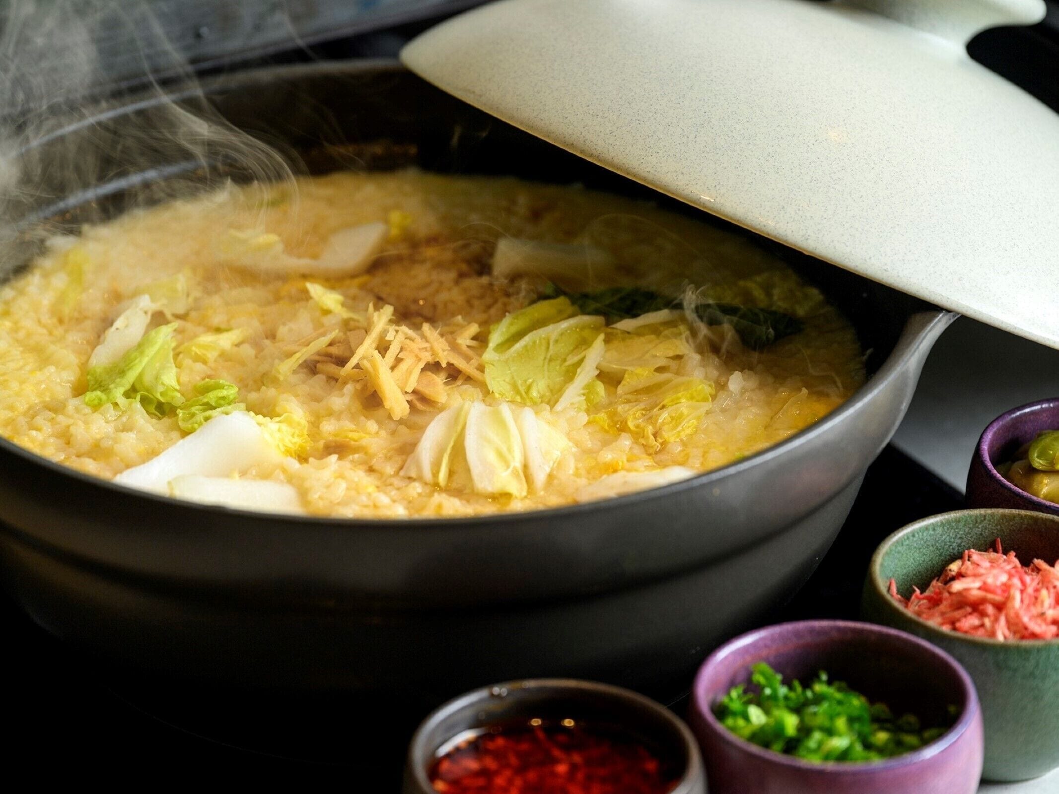 Una olla de sopa calenta amb diversos ingredients, vapor que s'escampa i petits bols de condiments al costat.