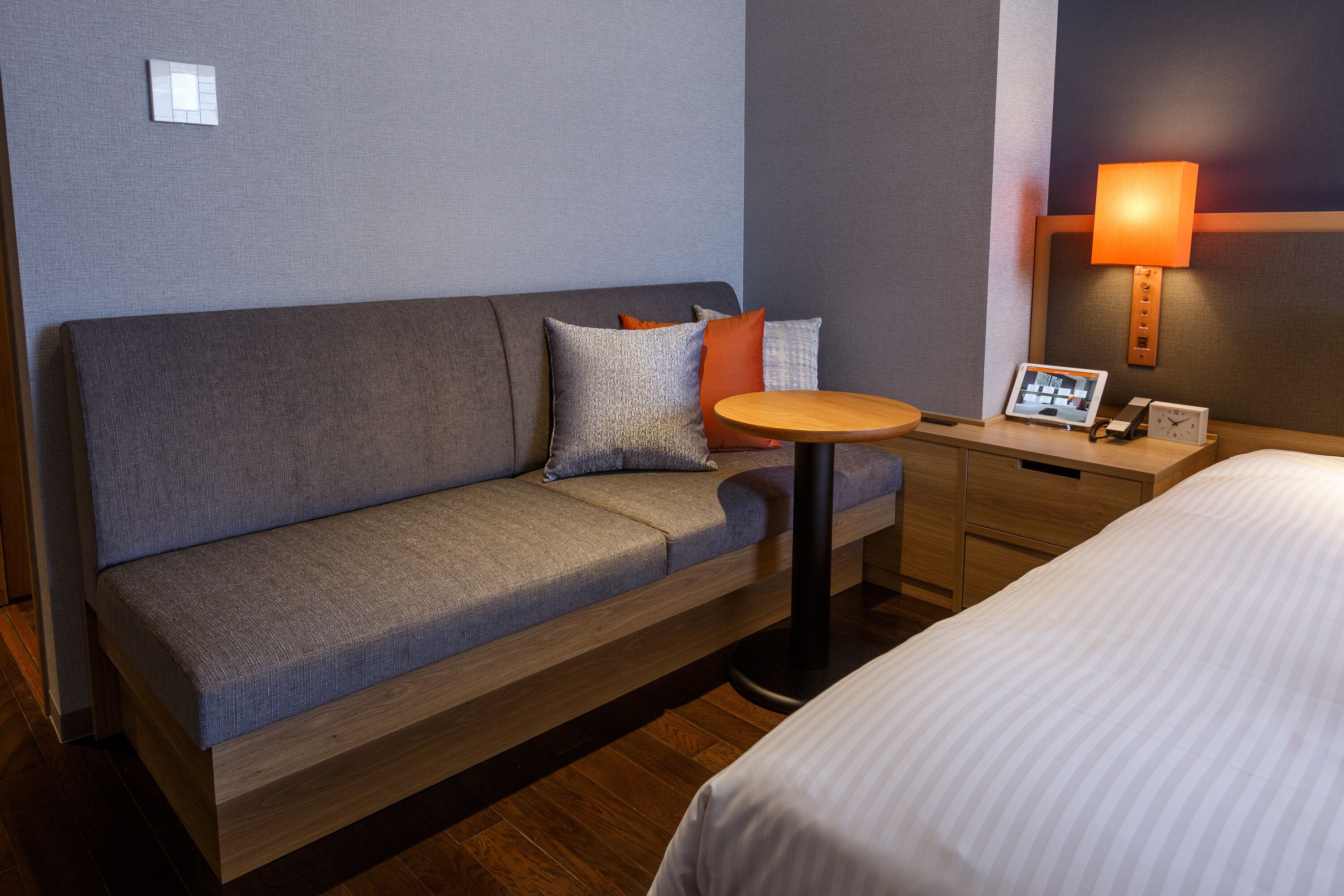 A hotel room with a grey sofa, a small round table, a bedside table with a lamp, and a bed.